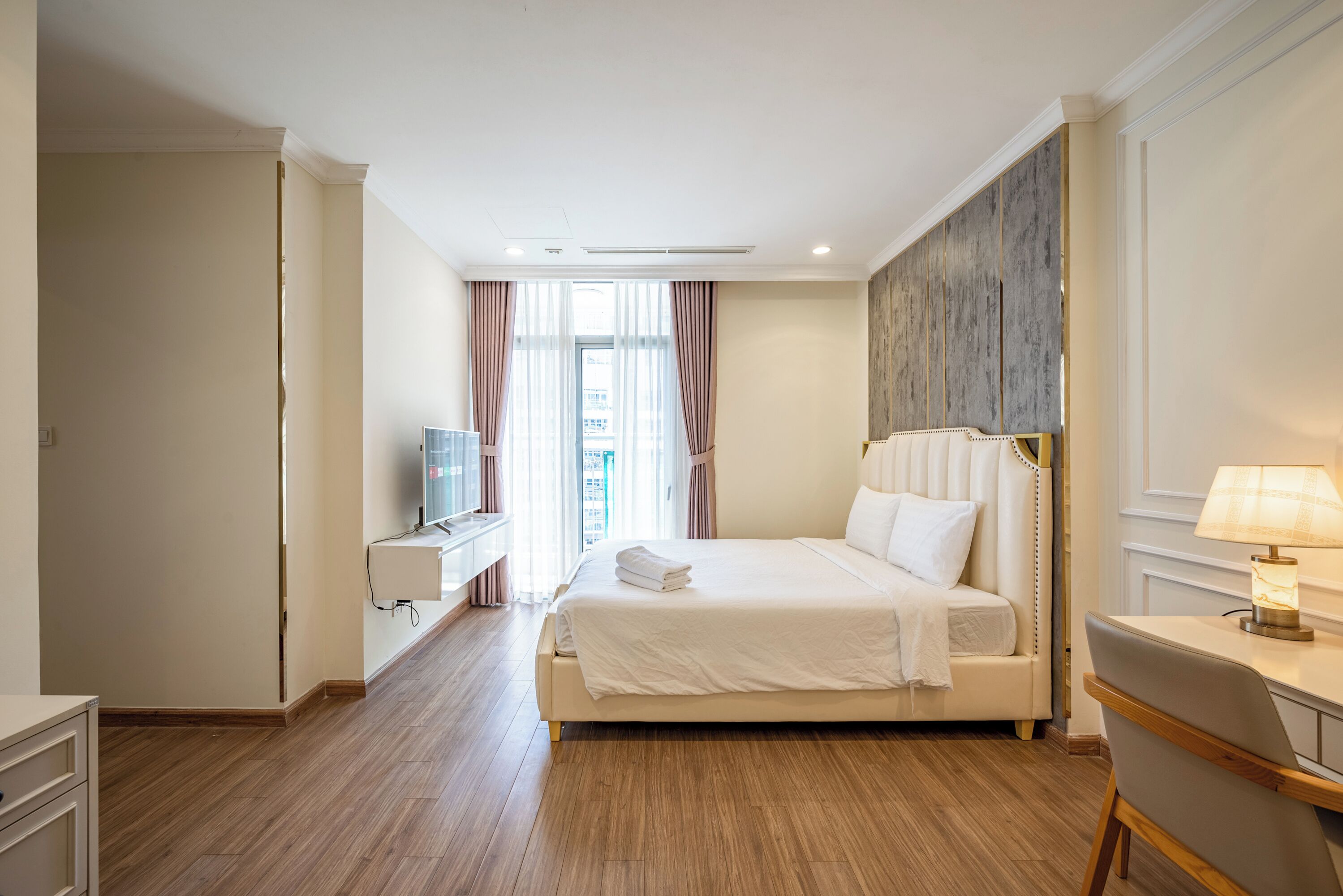 A hotel room with a large bed, a desk with a lamp, and a view of the city through the window.