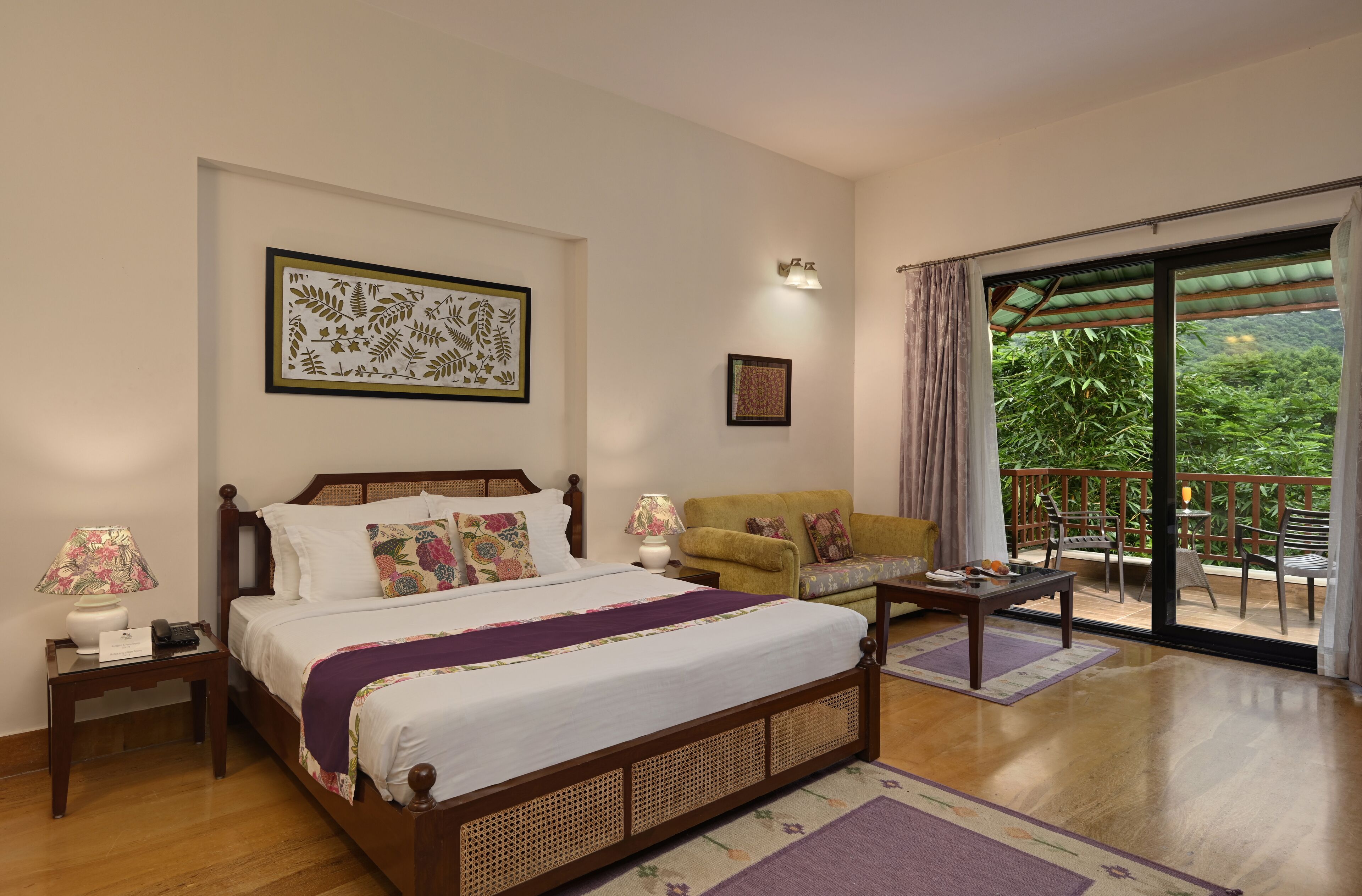 A bedroom with a large bed, a sofa, a table, and a balcony with a view of greenery.