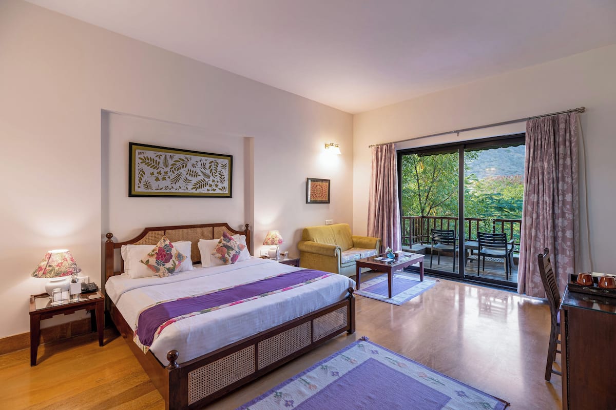A bedroom with a large bed, a nightstand, a chair, a table, and a balcony with a view of greenery.