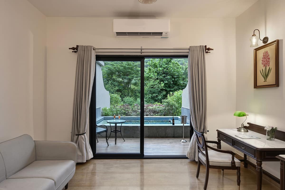 A room with a sofa, a desk with a lamp, a chair, and a view of a garden through a sliding door.