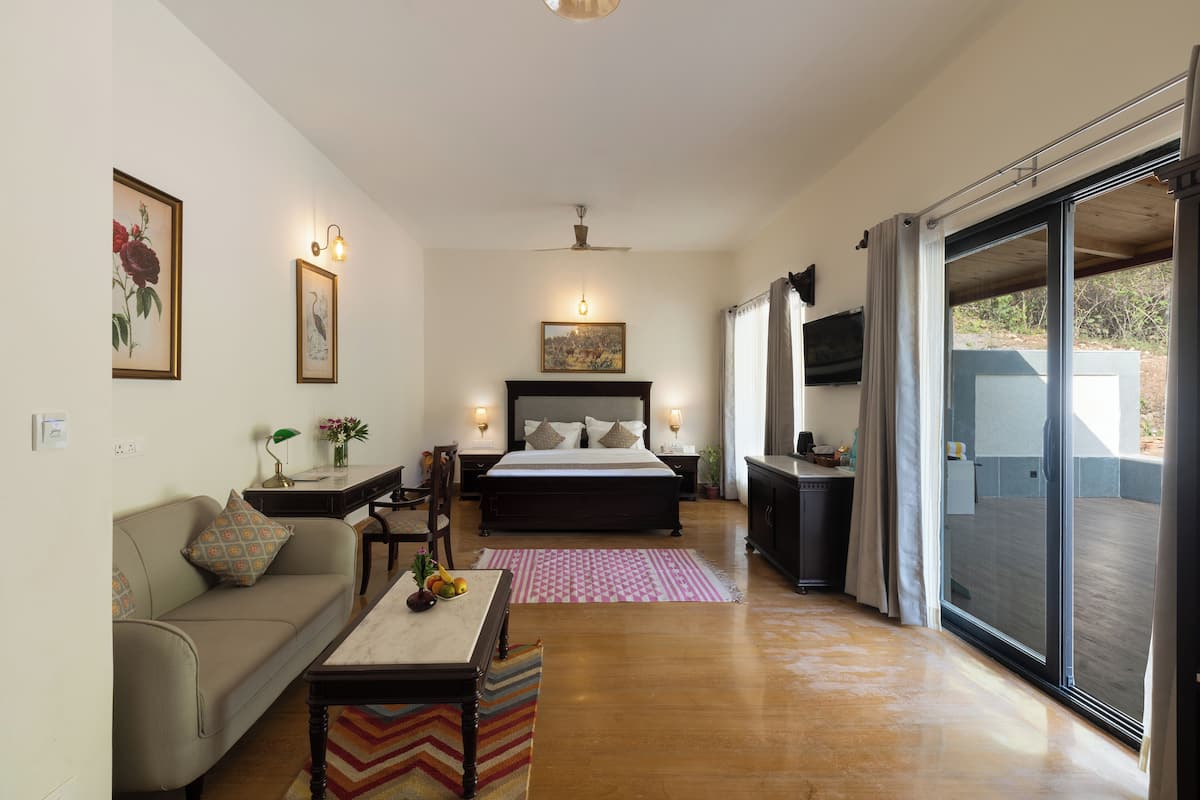A bedroom with a bed, a sofa, a dining table with fruits, a TV, and a sliding glass door to the outside.
