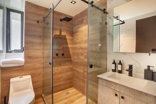 Un baño moderno con ducha envuelta en vidrio, paredes de madera, inodoro blanco y tocador de mármol con artículos de aseo.