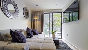 Een moderne woonkamer met een hoekbank, een ronde spiegel en een groot raam met uitzicht op bomen.