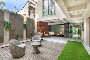 Un patio moderno al aire libre con mobiliario de mimbre, césped artificial verde y una puerta de cristal que da a una habitación con pared de madera y una silla colgante.
