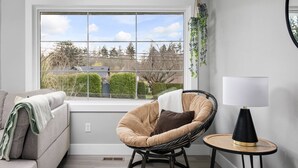 A modern living room with a grey sofa, a wicker chair, a small round table with a lamp, and a large window with a view of trees and a house. - Spectacular Single Level Estate, Pet Friendly, Huge Fully Fenced Backyard, Covered Patio, Game Room (Portland)