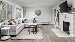 A modern living room with a grey sofa, a round coffee table, a white fireplace, and a large window with a view of trees. - Spectacular Single Level Estate, Pet Friendly, Huge Fully Fenced Backyard, Covered Patio, Game Room (Portland)