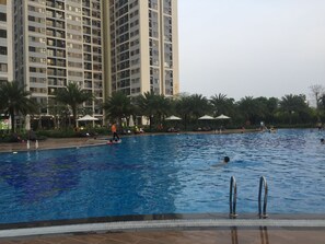 A swimming pool with people swimming and relaxing, surrounded by palm trees and a tall building in the background. - Vinhome Grand Park Homestay House (Thành phố Hồ Chí Minh)