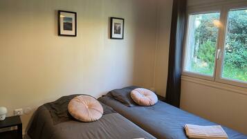 Une chambre avec deux lits, chacun muni d’un oreiller et d’une serviette pliée. Les murs sont ornés de tableaux encadrés, il y a une table de chevet avec une lampe, et une fenêtre donnant sur un paysage verdoyant.