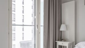 Une chambre d’hôtel avec un lit, une table de chevet avec une lampe, une grande fenêtre avec des rideaux et une vue sur un bâtiment à l’extérieur.