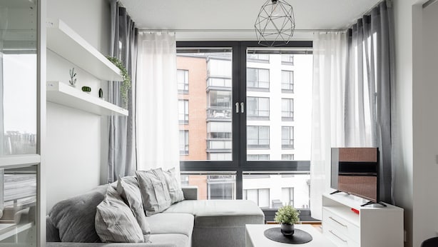 Un salon moderne avec un canapé d’angle gris, une table basse blanche et un téléviseur à  écran plat posé sur un meuble blanc. On y trouve des étagères blanches ornées d’objets décoratifs et une grande fenêtre avec des rideaux.