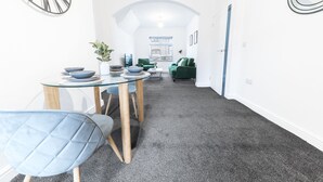 Un intérieur moderne avec un coin repas comprenant une table et des chaises en verre, un canapé vert et un couloir menant à une porte.