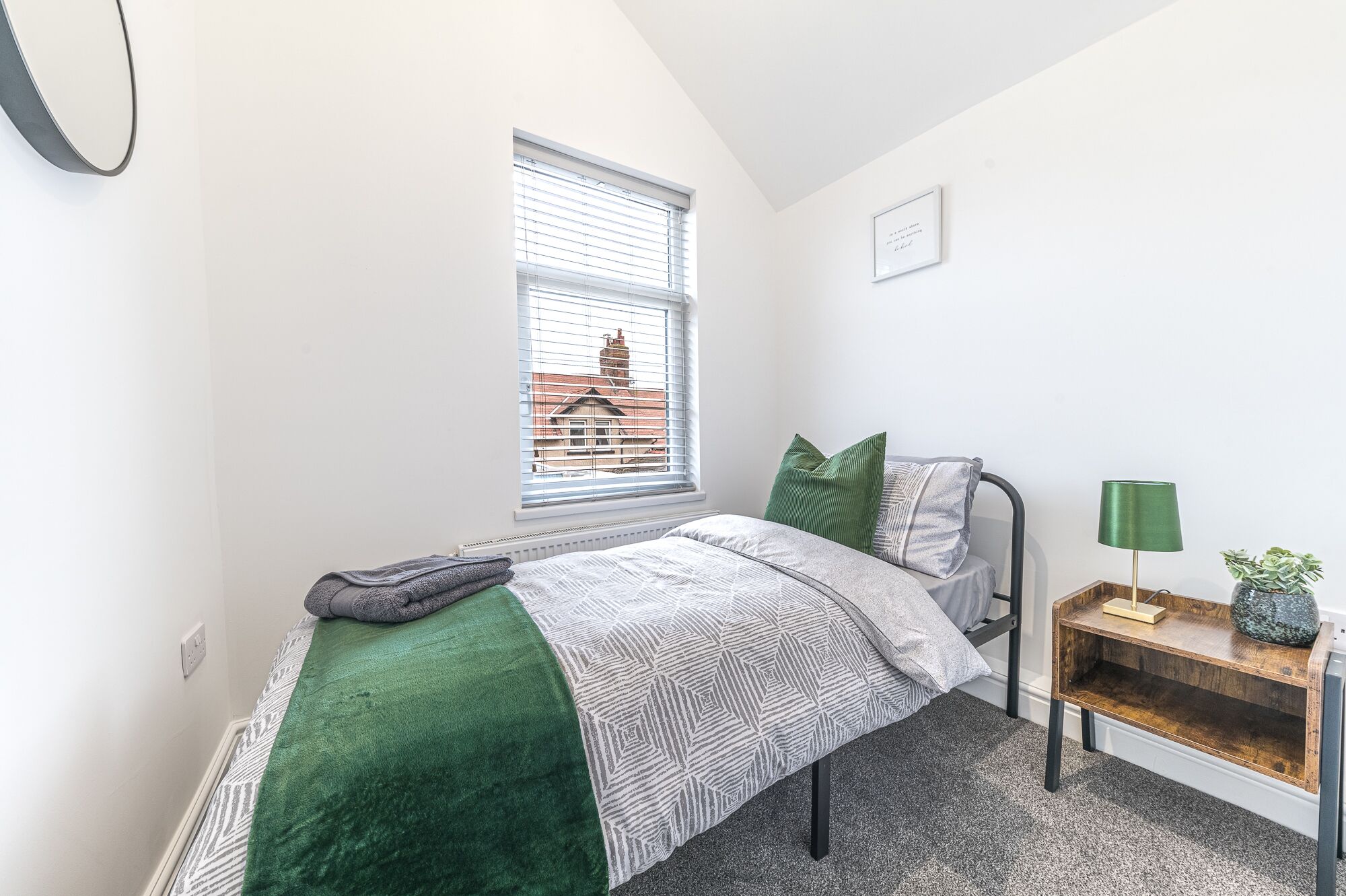 Une chambre à coucher avec un lit, une table de chevet avec une lampe, une fenêtre avec des stores et une vue sur les bâtiments à l’extérieur.