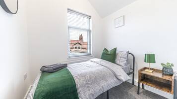 Une chambre à coucher avec un lit, une table de chevet avec une lampe, une fenêtre avec des stores et une vue sur les bâtiments à l’extérieur.
