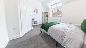 Une chambre à coucher avec un lit, une fenêtre avec des stores, un radiateur, un porte-manteau et une porte.