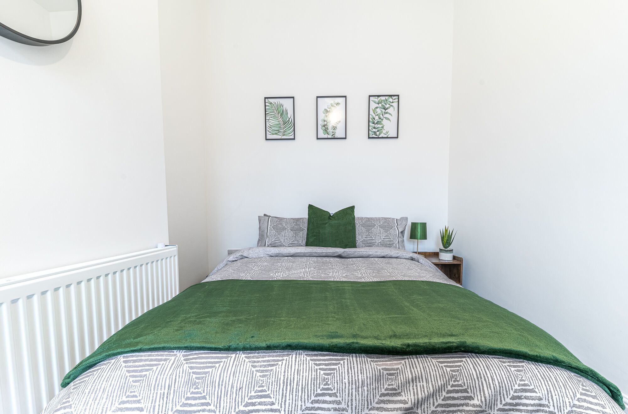 Une chambre à coucher comprenant un lit, une table de chevet, une lampe et trois reproductions botaniques encadrées accrochées au mur.