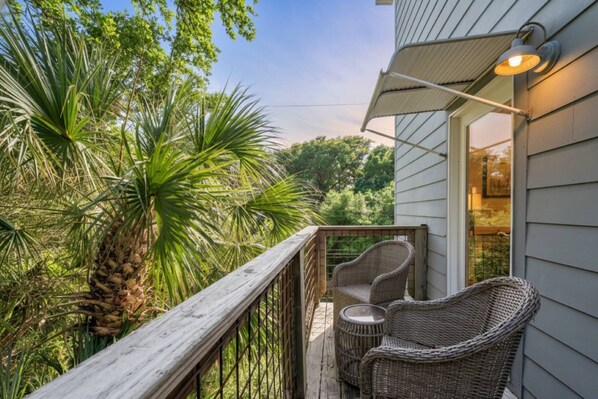 A wooden deck with wicker furniture, a palm tree, and a house with a sloped roof and a light fixture.