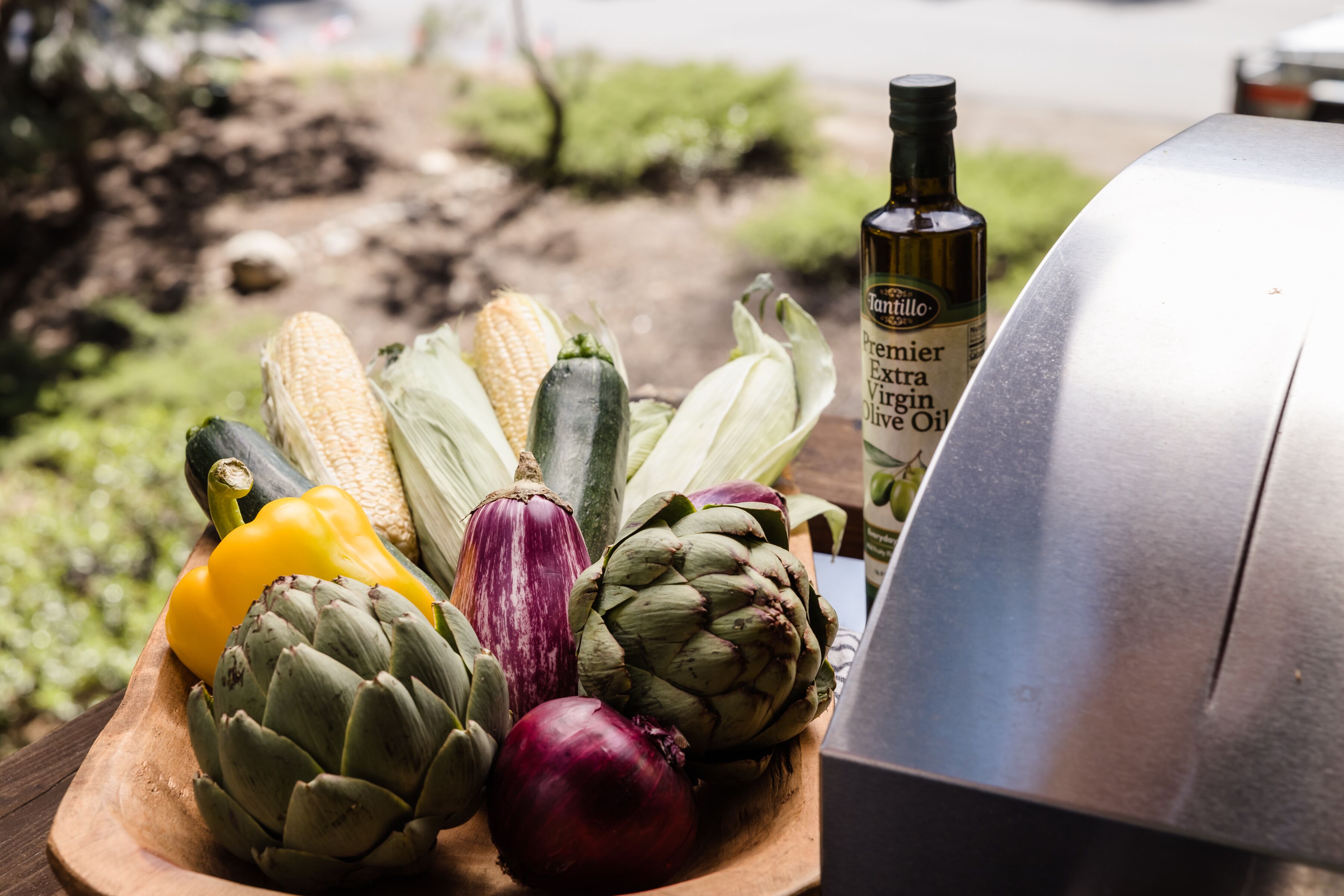 A wooden board with various vegetables and a bottle of extra virgin olive oil.