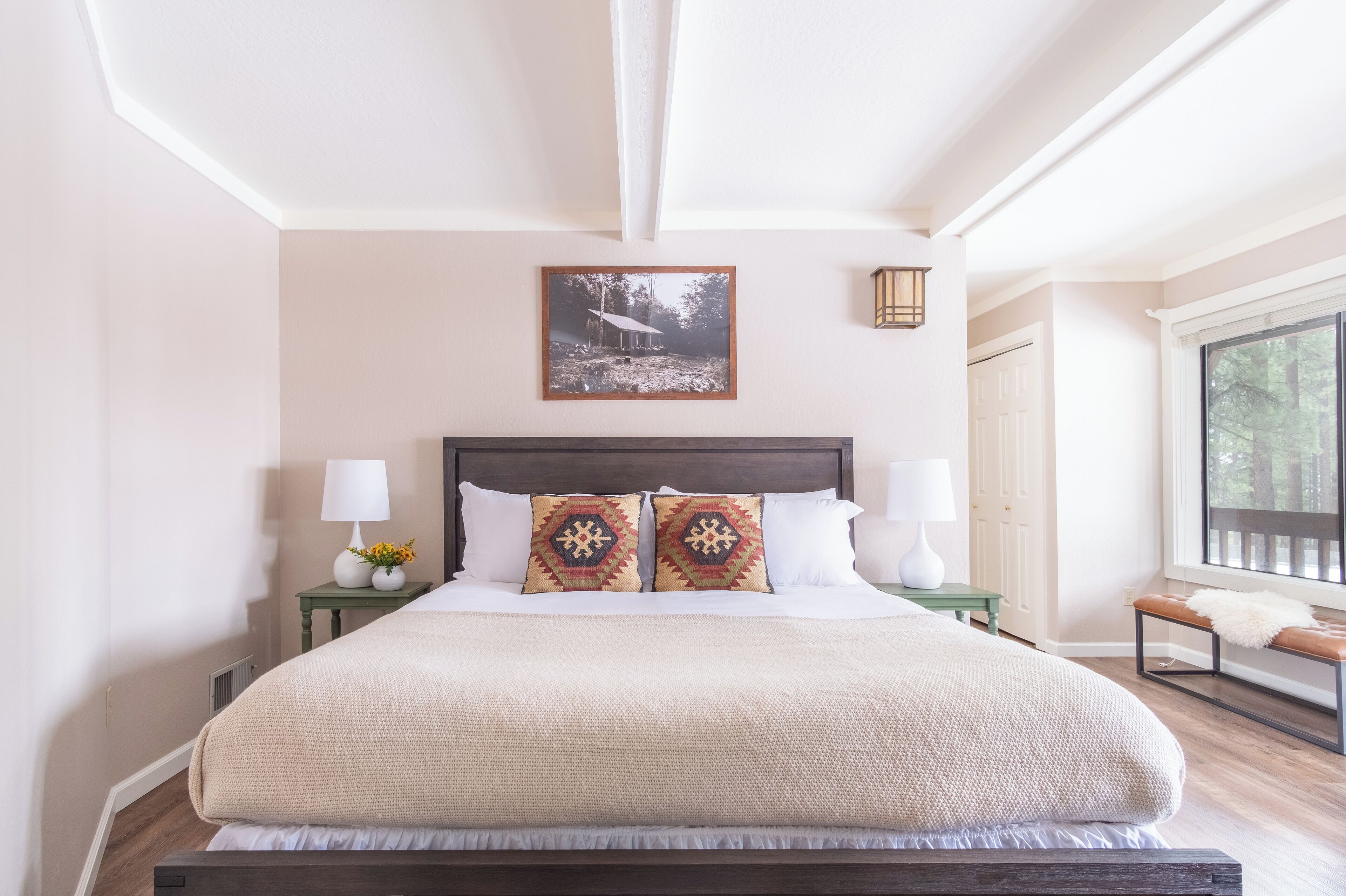 A well-lit bedroom with a large bed, two nightstands, a bench, and a window with a view of trees.