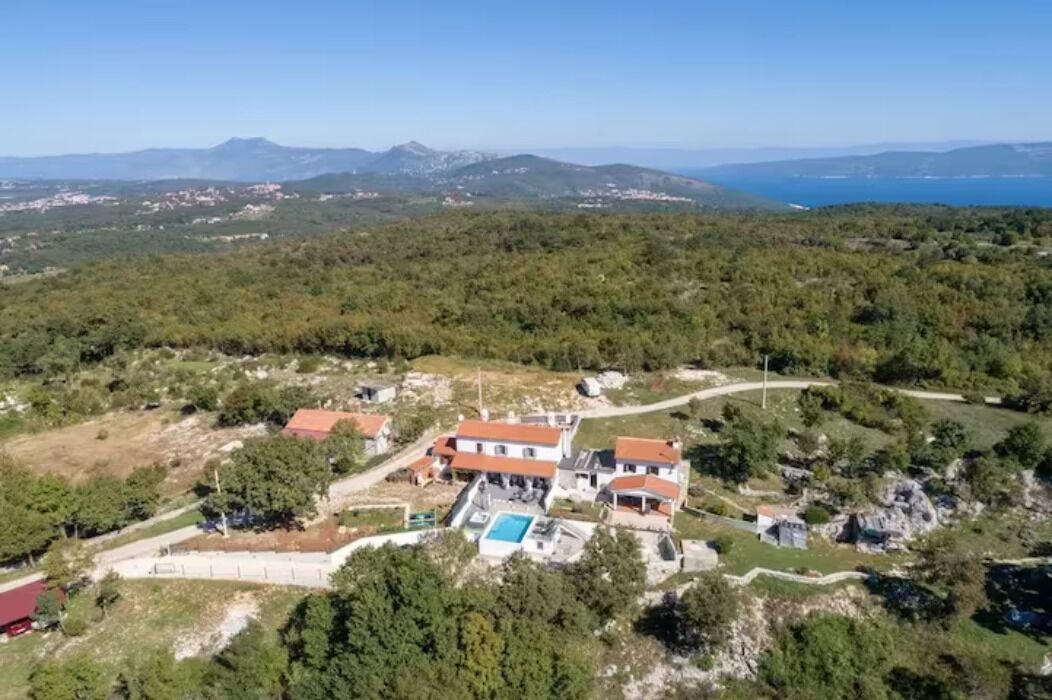 A residential area with houses, a swimming pool, and a clear view of the sea and mountains in the background.