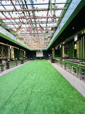 A spacious indoor area with artificial grass flooring, a glass ceiling with hanging decorations, and green walls with vertical gardens.