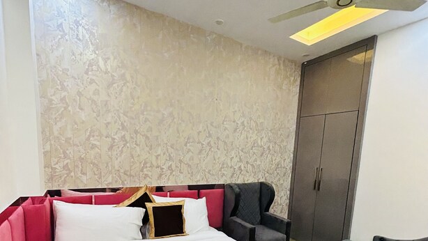 A modern bedroom with a large bed, a grey armchair, a small table, and a wardrobe. - Shree Jee Hotel (Faridabad)