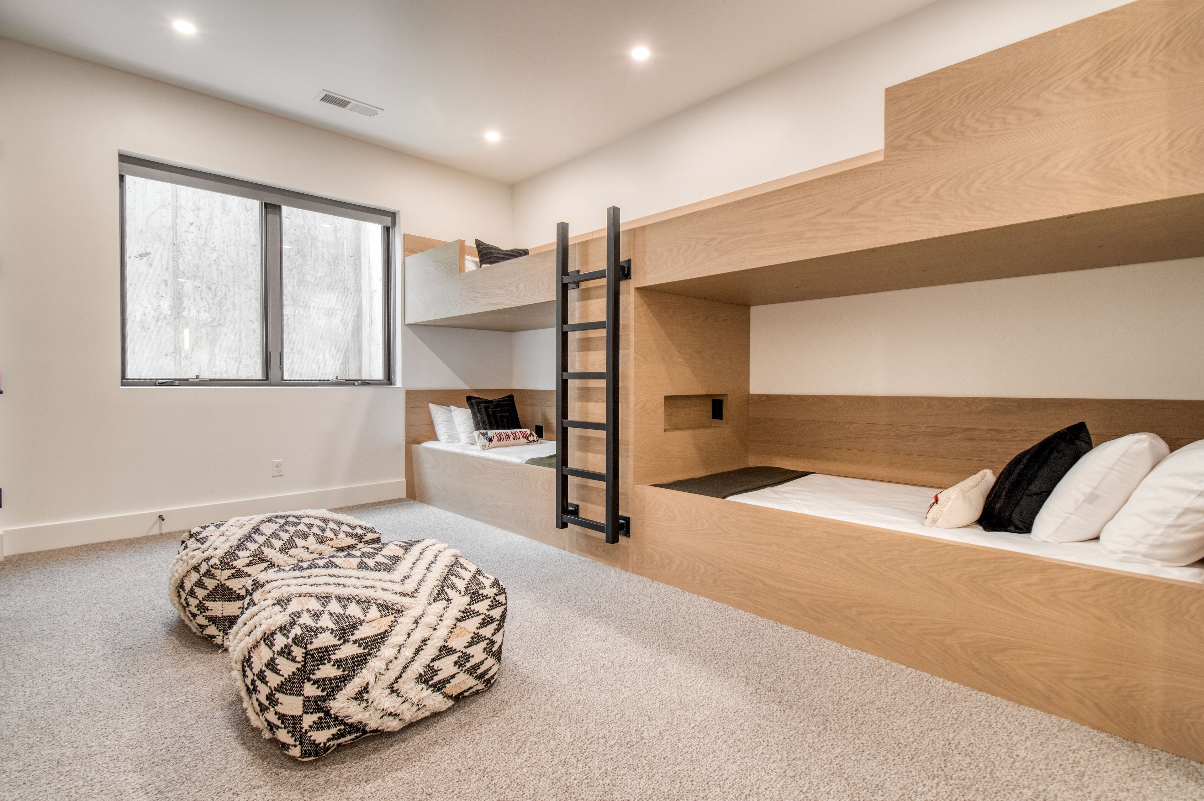 A bunk bed room with a ladder, two ottomans, and a large window.