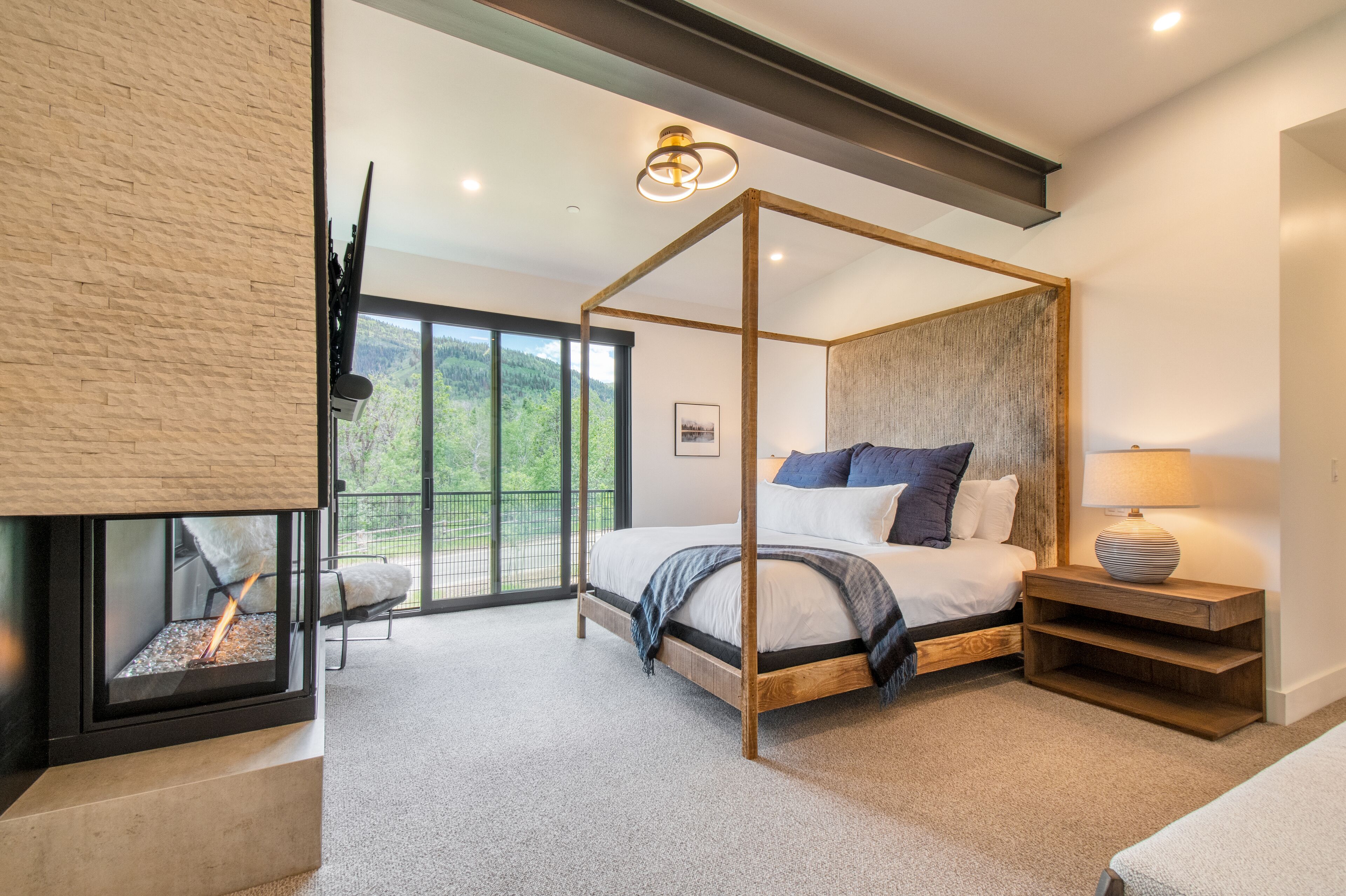 A modern bedroom with a four-poster bed, a fireplace, and a large window offering a view of the outdoors.