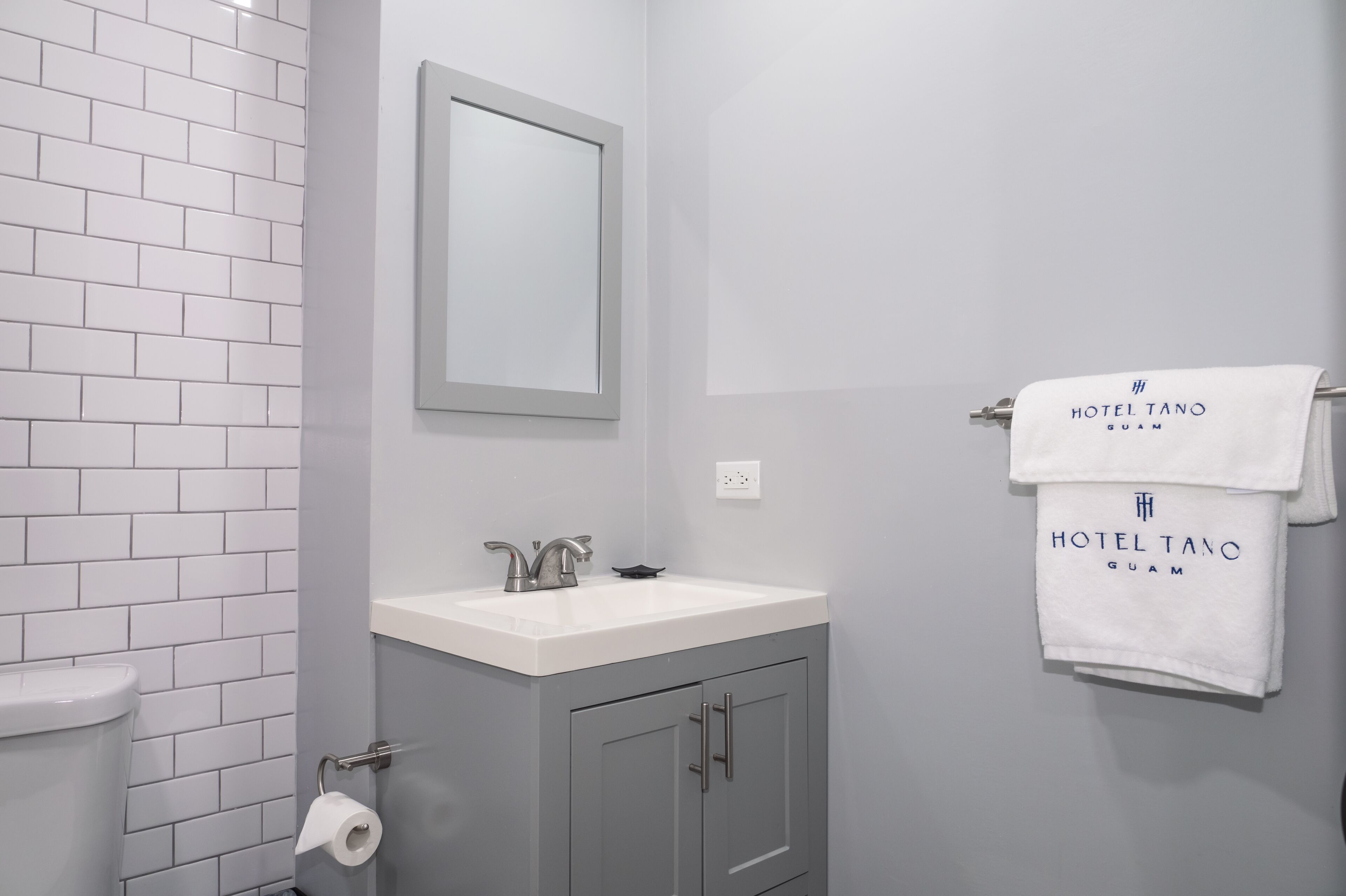 A bathroom with a white sink, a mirror, and a towel rack with a towel labeled "HOTEL TANO GUAM".