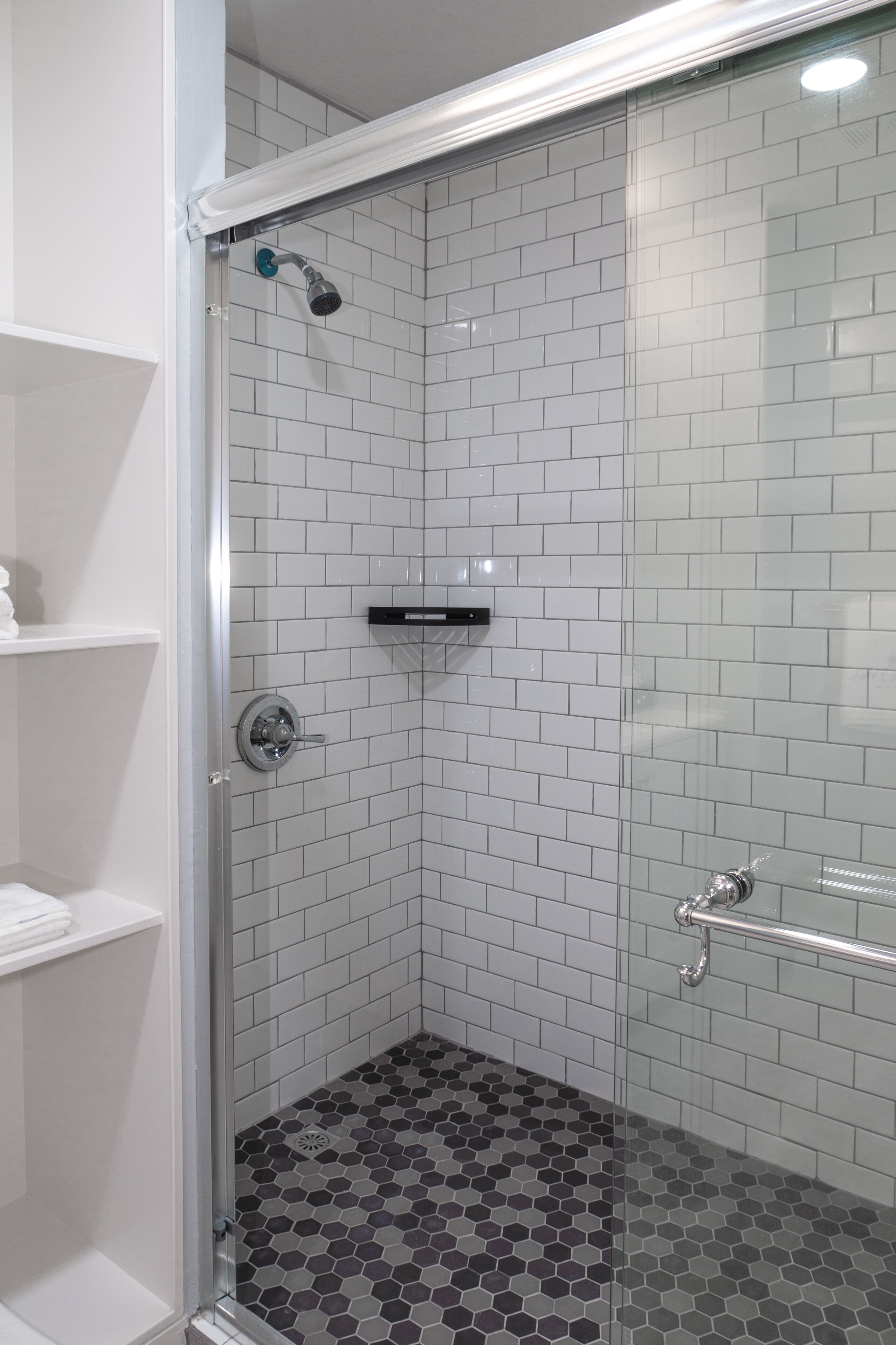 Ducha con cerramiento de vidrio, paredes de azulejos blancos y piso con diseño hexagonal.