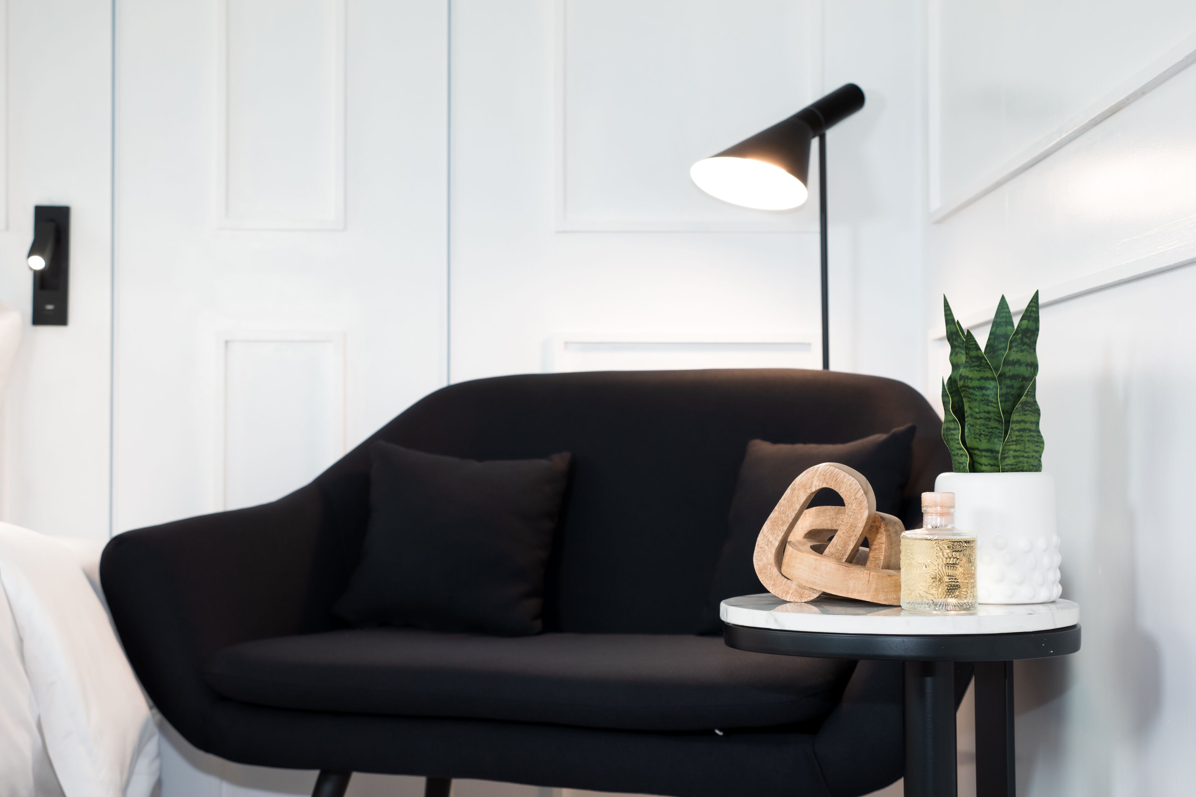 A modern living room with a black sofa, a small round table with a lamp, a potted plant, and a decorative item.