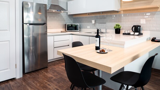 A modern kitchen with a stainless steel refrigerator, a microwave, a coffee maker, and a wooden dining table with black chairs. - HOTEL TANO (Tamuning)