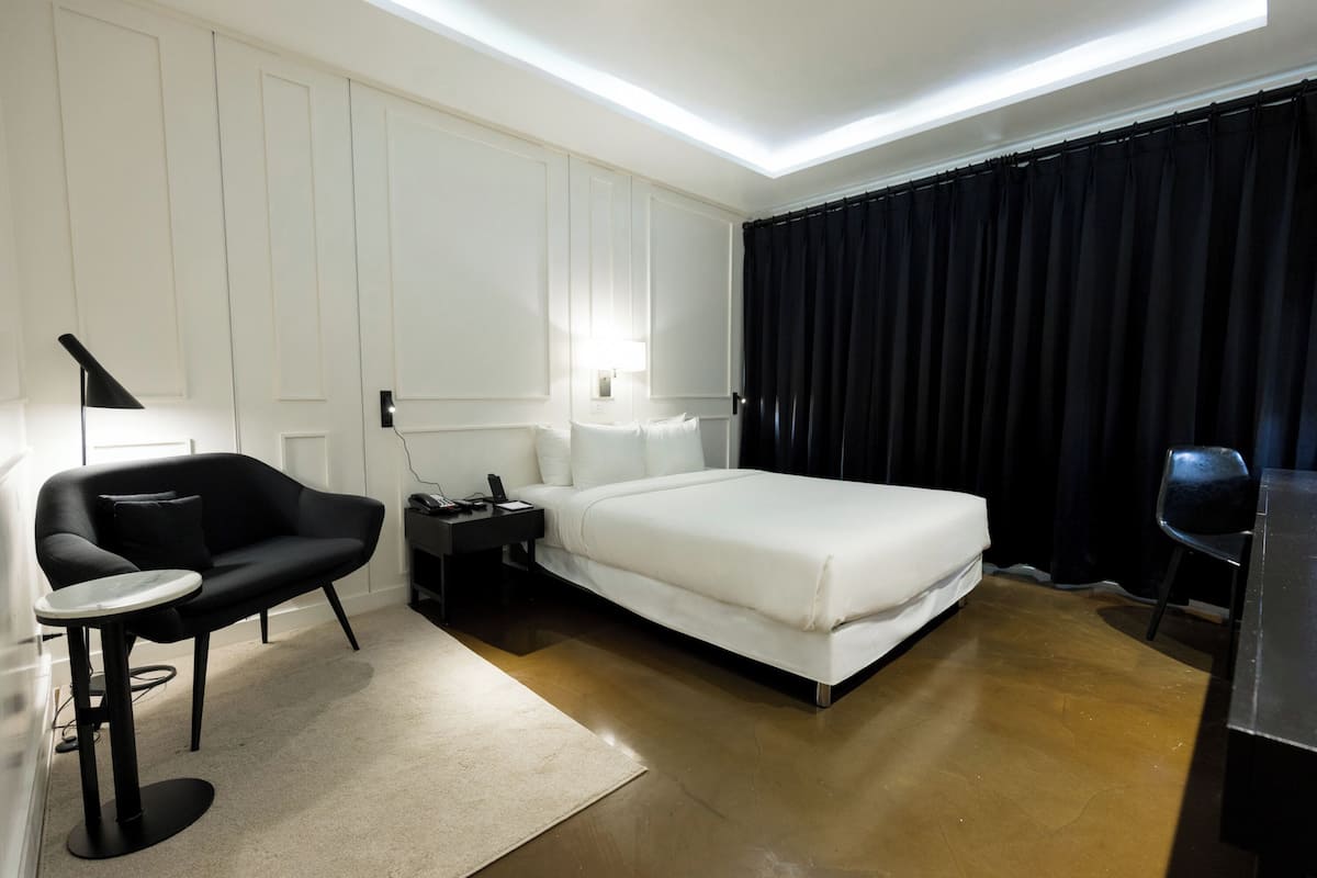 A modern hotel room with a bed, a desk, a chair, a lamp, and a nightstand.