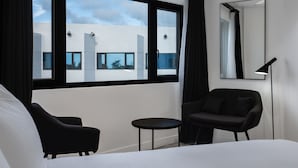 A hotel room with a large window, a bed, a chair, a small table, and a floor lamp. - HOTEL TANO (Tamuning)