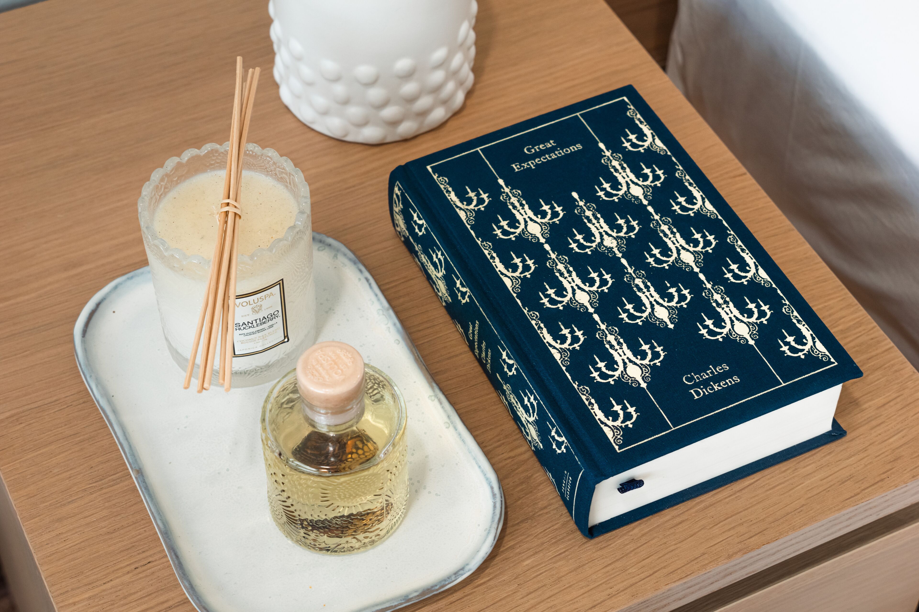 Una bandeja con un vaso de vela perfumada, un frasco de colonia y un libro titulado 'Grandes Esperanzas' de Charles Dickens.