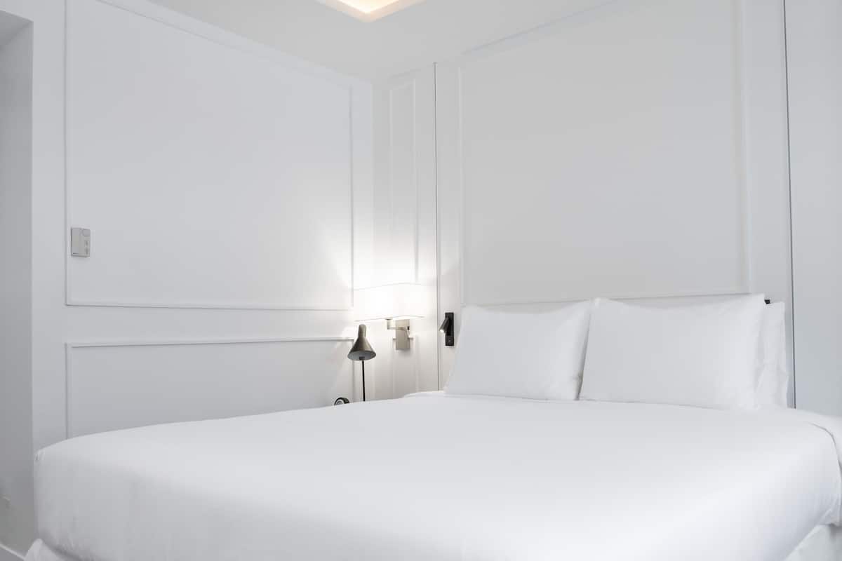 A white, minimalist bedroom with a large bed, a bedside table, and a wall-mounted light fixture.