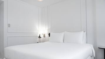 Un dormitorio blanco y minimalista con una cama grande, una mesita de noche y un aplique de pared.