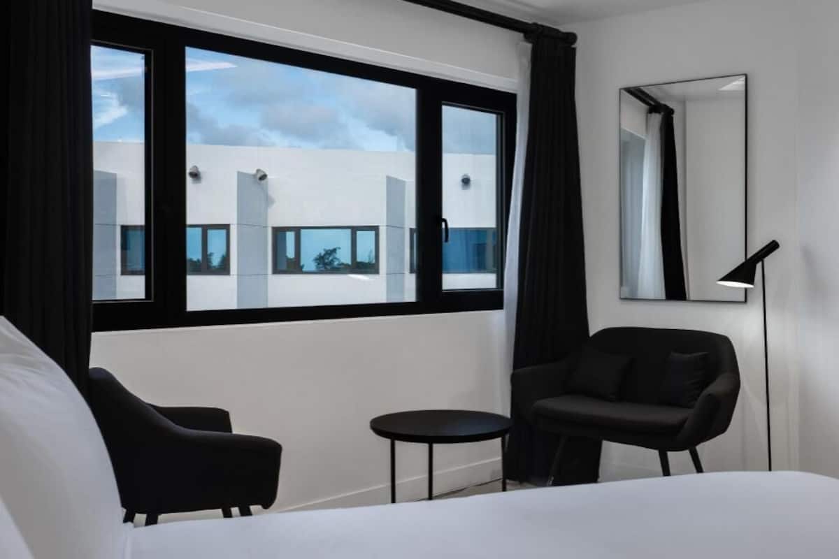 A modern hotel room with a large bed, two black chairs, a small round table, a large window with black frames, and a mirror on the wall.