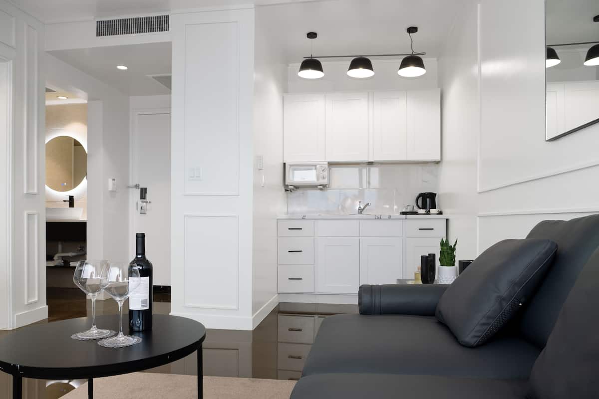A modern living room with a grey sofa, a black coffee table with wine glasses and a bottle, and a kitchen area with white cabinets and a microwave.