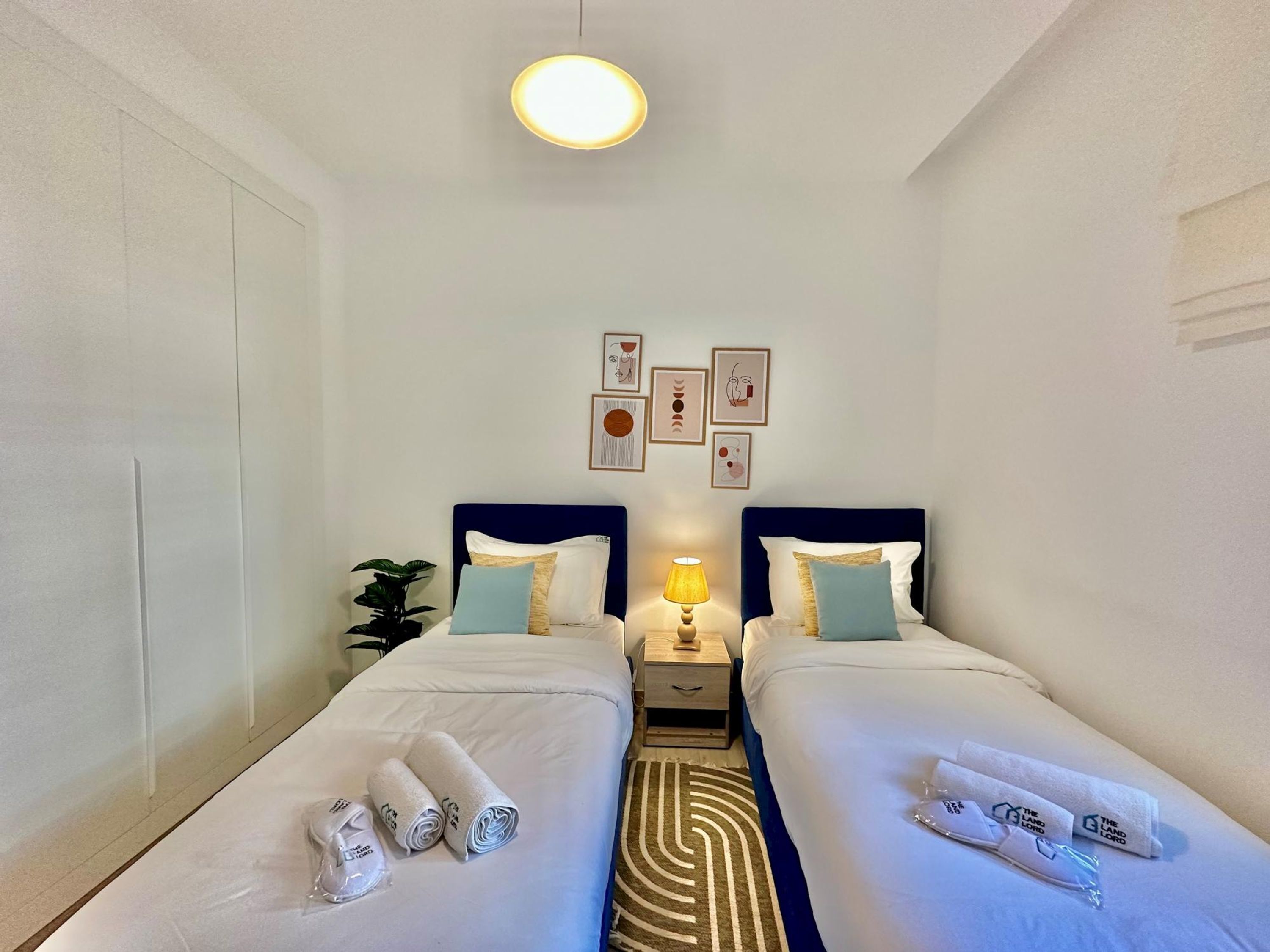 Een hotelkamer met twee bedden, een nachtkastje met een lamp en een muur met ingelijste kunstwerken.