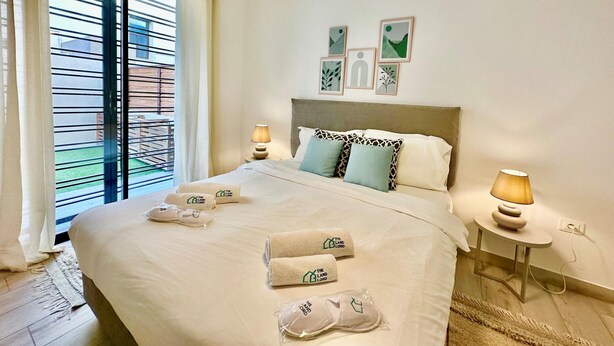 A hotel room with a bed, two bedside lamps, a window with blinds, and artwork on the wall. - Rd Jade apt Neuf Avec Jardin Proche des Commodit s (La Marsa)