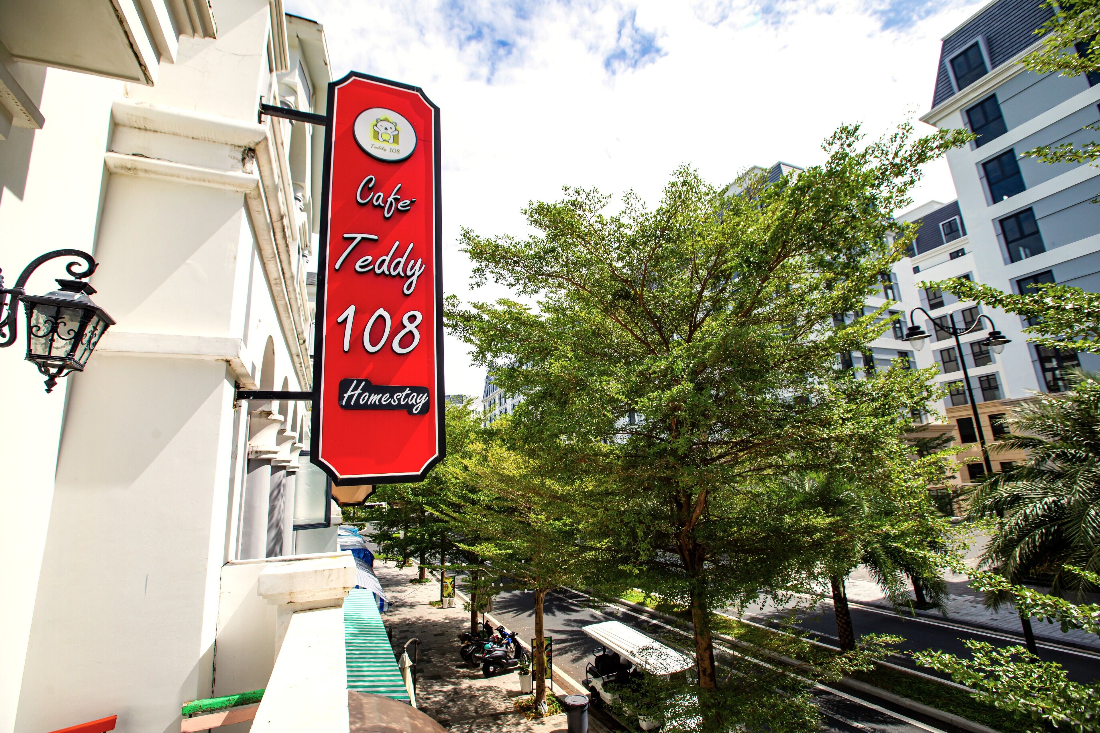 Punainen kyltti, jossa lukee ”Cafe Teddy 108”, ja sen yhteydessä oleva ”HomeStay”-logo, on kiinnitetty valkoiseksi maalattuun rakennukseen, jonka yläpuolella on vihreä markiisi.