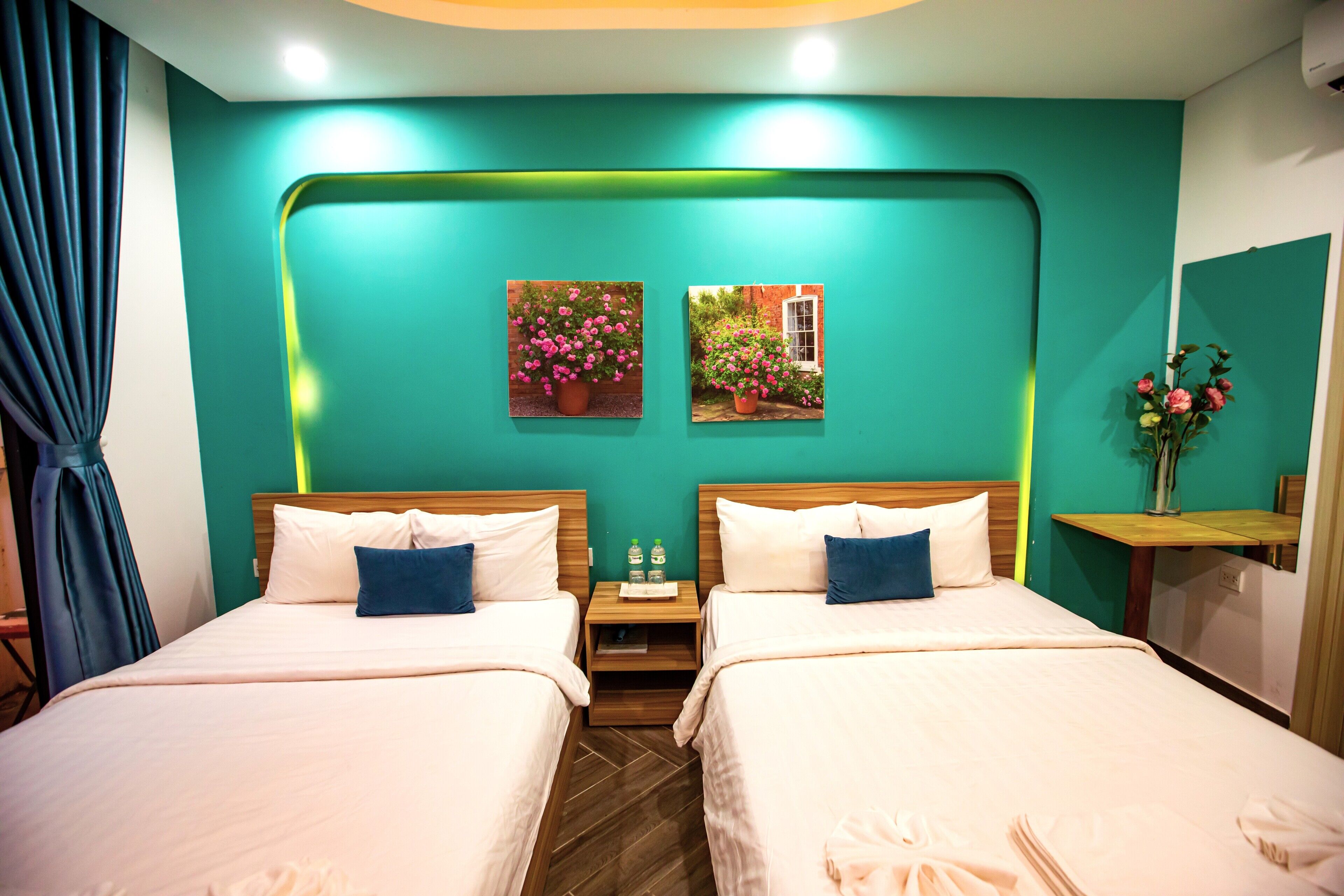 Ein Hotelzimmer mit zwei Betten, türkisfarbenen Wänden und einem kleinen Tisch mit Blumen in einer Vase.