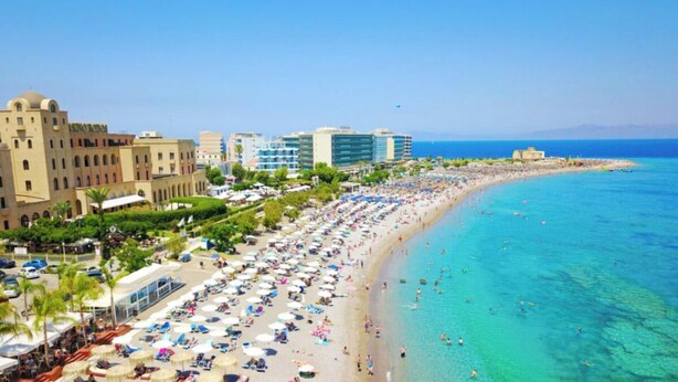 A beach with numerous umbrellas and sun loungers, a clear blue sea, and buildings in the background. - Elli Down Town Apartment Karma (Rodos)