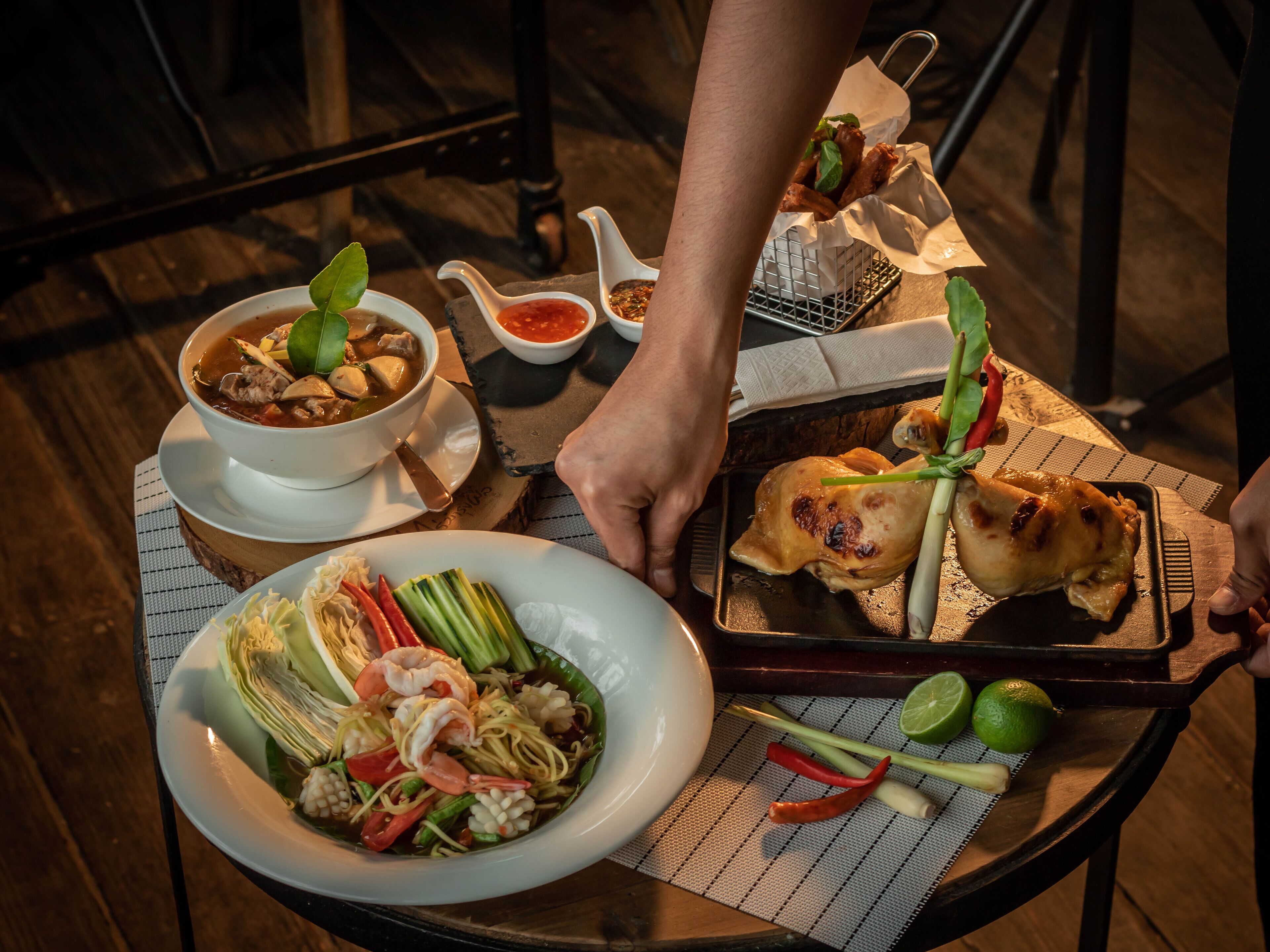 A table with a variety of dishes, including a bowl of soup, a plate of noodles with shrimp, and grilled chicken. There are also condiments and fresh herbs on the table.