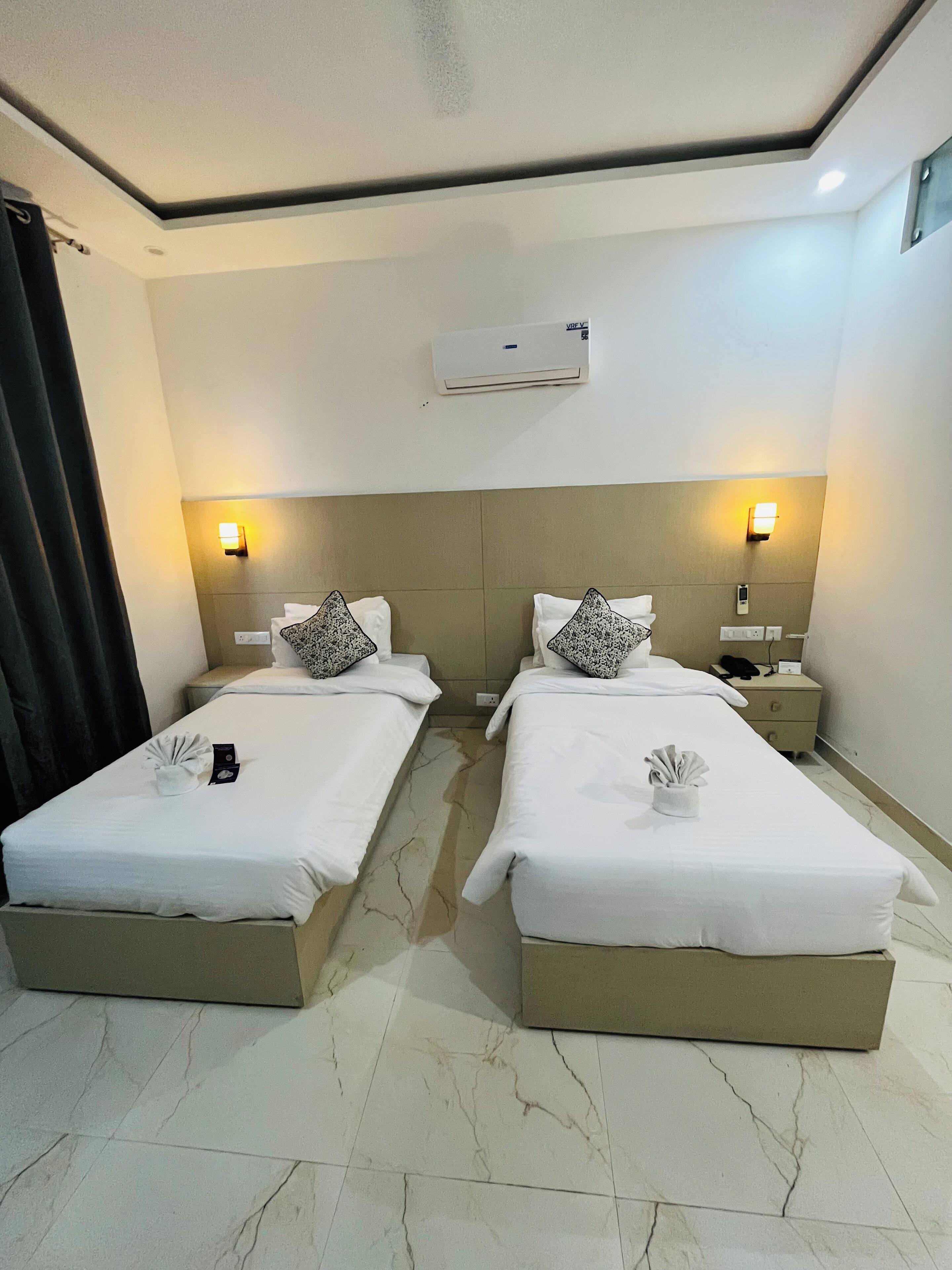 A hotel room with two beds, each with a pillow and a folded towel. There is a nightstand with a phone and a lamp.