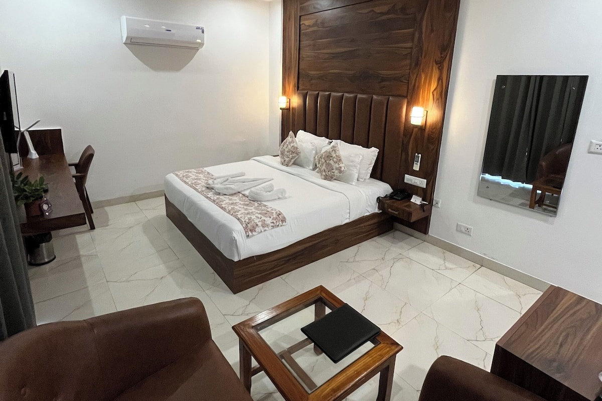 A modern hotel room with a large bed, two nightstands, a wooden headboard, a ceiling fan, an air conditioning unit, a small table, two brown leather chairs, and a mirror on the wall.
