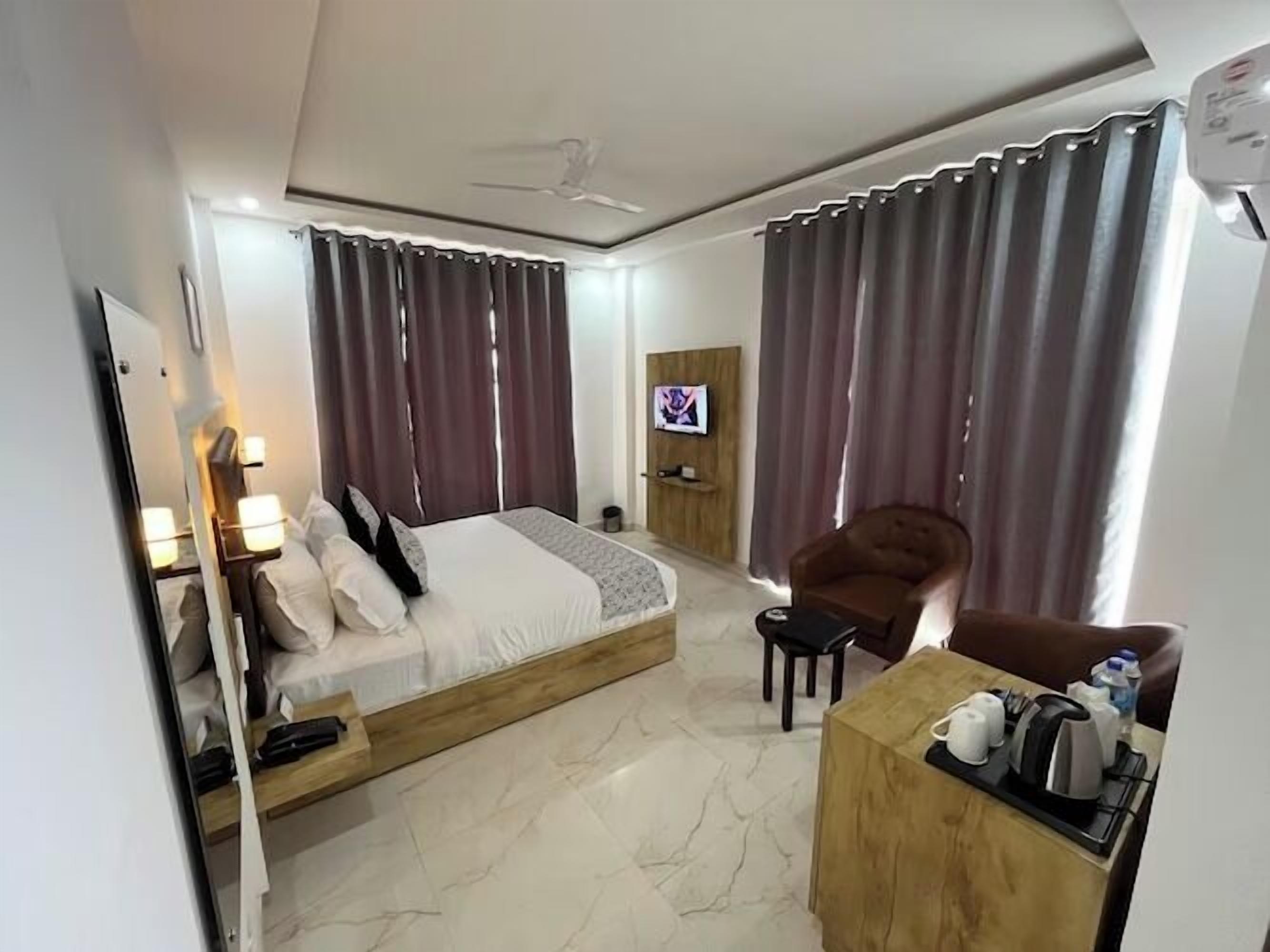 A hotel room with a bed, a desk with a kettle, a chair, a TV, and curtains.