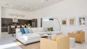 Uma sala de estar moderna com um sofá, poltronas, uma mesa de centro e uma área de cozinha ao fundo.