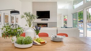 Uma sala de estar moderna com televisão de tela plana, lareira e área de jantar com mesa de madeira, cadeiras e uma tábua de frutas e queijos.