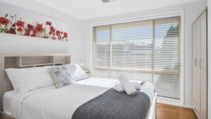 Un dormitorio moderno con una cama grande, ropa de cama blanca, un plaid gris y un cuadro de flores rojas.